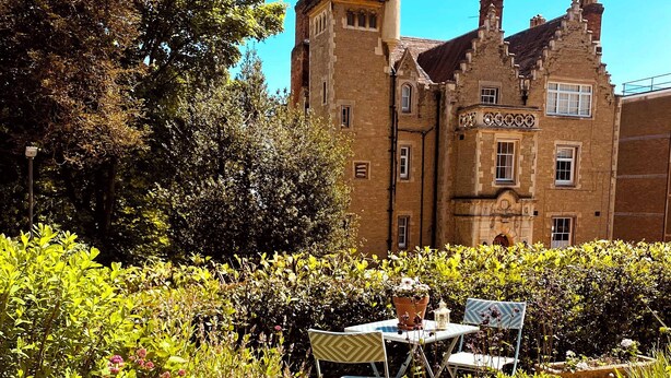 A historic building with a tower, surrounded by lush greenery and a small outdoor seating area with a table and chairs. - The Escape (Malvern)
