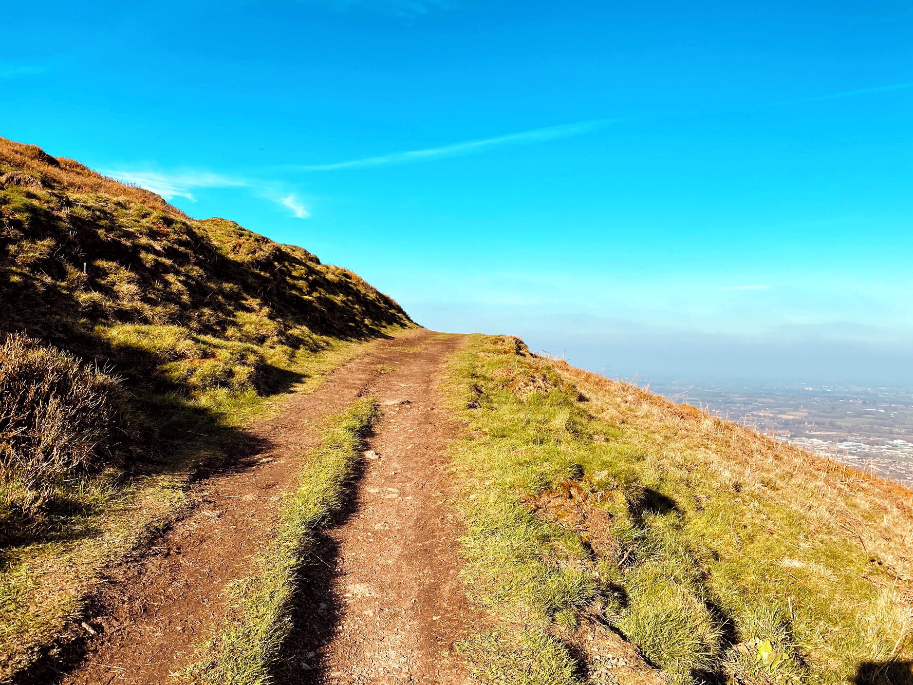 A dirt path on a hillside with a clear view of a distant cityscape.