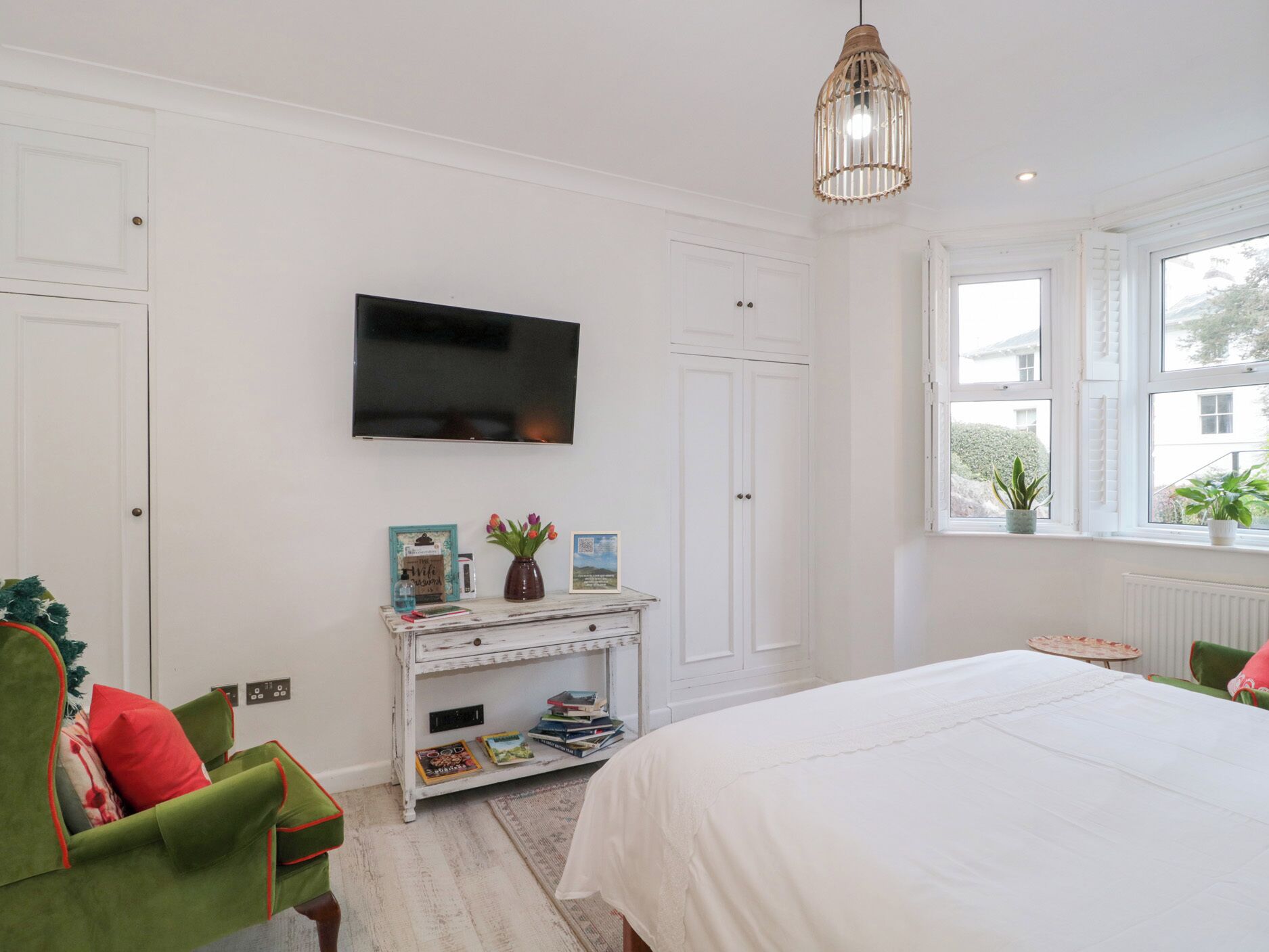Une chambre avec un lit, un téléviseur fixé au mur, une petite table console blanche ornée de livres et d’un vase, et une fenêtre avec des plantes.