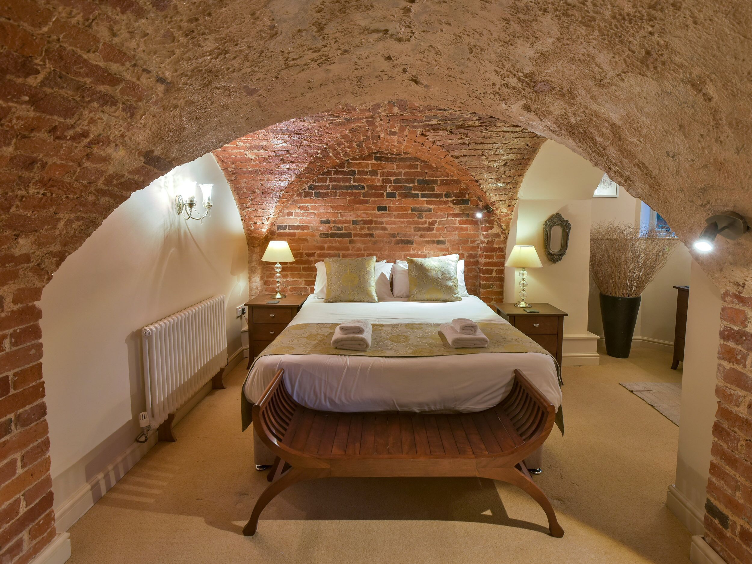 A cozy room with a brick wall, a wooden bench, a bed with white linens, and a potted plant.