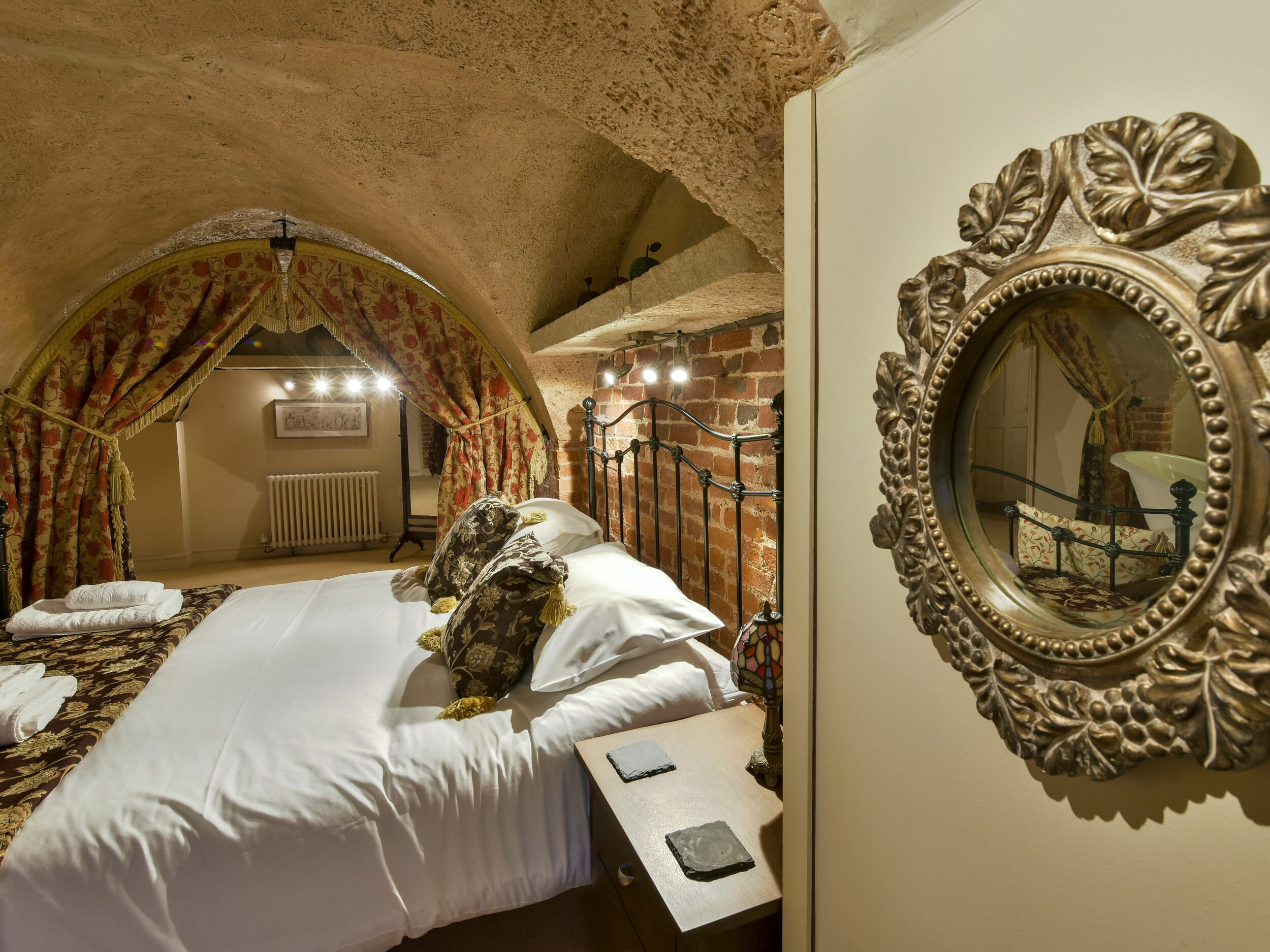 A bedroom with a four-poster bed, a large ornate mirror, and a brick wall.