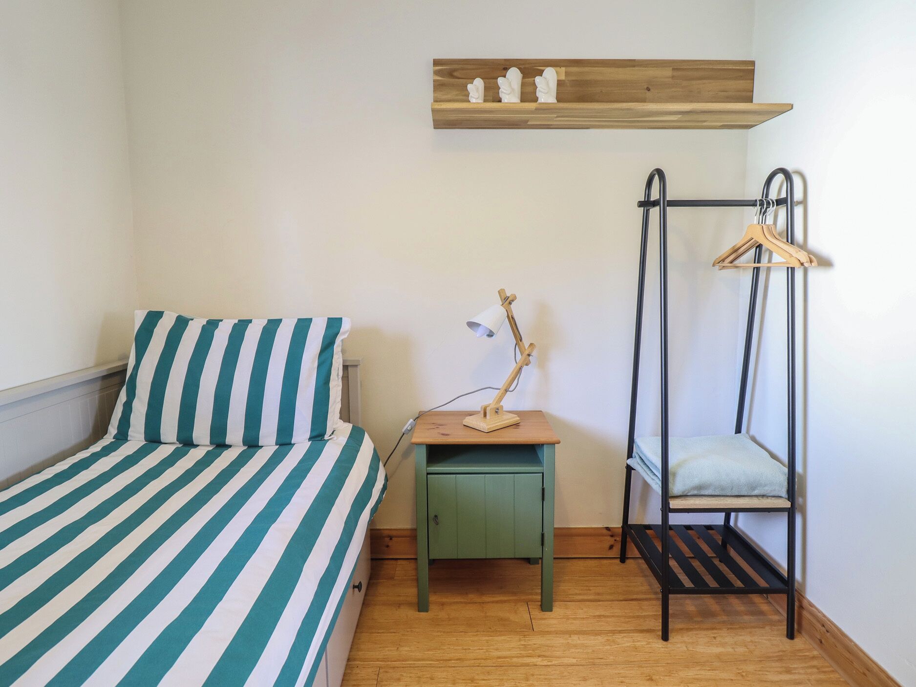A single bed with striped bedding, a small bedside table with a lamp, a hanging chair, and a shelf with decorative items.