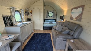 Un intérieur compact et chaleureux comprenant un coin cuisine, un lit, un canapé et une fenêtre ronde.