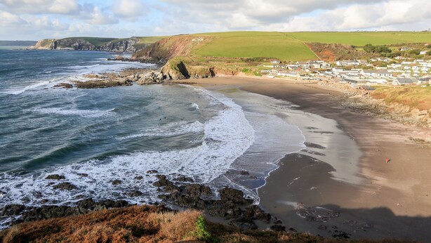 A coastal scene with a sandy beach, rocky shoreline, and a village in the background. - 2 The Bay (Kingsbridge)