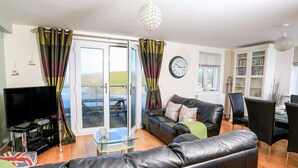 A living room with a leather sofa, a glass coffee table, a flat-screen TV, and a dining area with chairs and a table. - 2 The Bay (Kingsbridge)