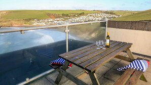 A wooden picnic table with a bottle of wine and glasses on a balcony overlooking a village. - 2 The Bay (Kingsbridge)