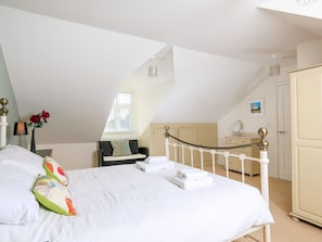 A bedroom with a sloped ceiling, a bed with white linens, a bedside table, a lamp, a vase with flowers, a window with blinds, a chair, a dresser, and a door. - 2 The Bay (Kingsbridge)