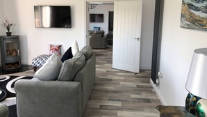 A modern living room with a grey sofa, a flat-screen TV, a fireplace, and a dining area in the background. - 1 Barn Cottages (Whitchurch)