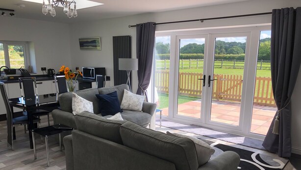 A modern living room with a grey sofa, a dining area with a glass table, and a view of a green landscape through large sliding doors. - 1 Barn Cottages (Whitchurch)