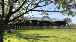 A grassy yard with a wooden fence, a tree with white blossoms, and a building with solar panels. - 1 Barn Cottages (Whitchurch)