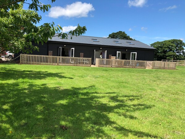 A building with a dark exterior, multiple doors, and a fenced yard. - 1 Barn Cottages (Whitchurch)