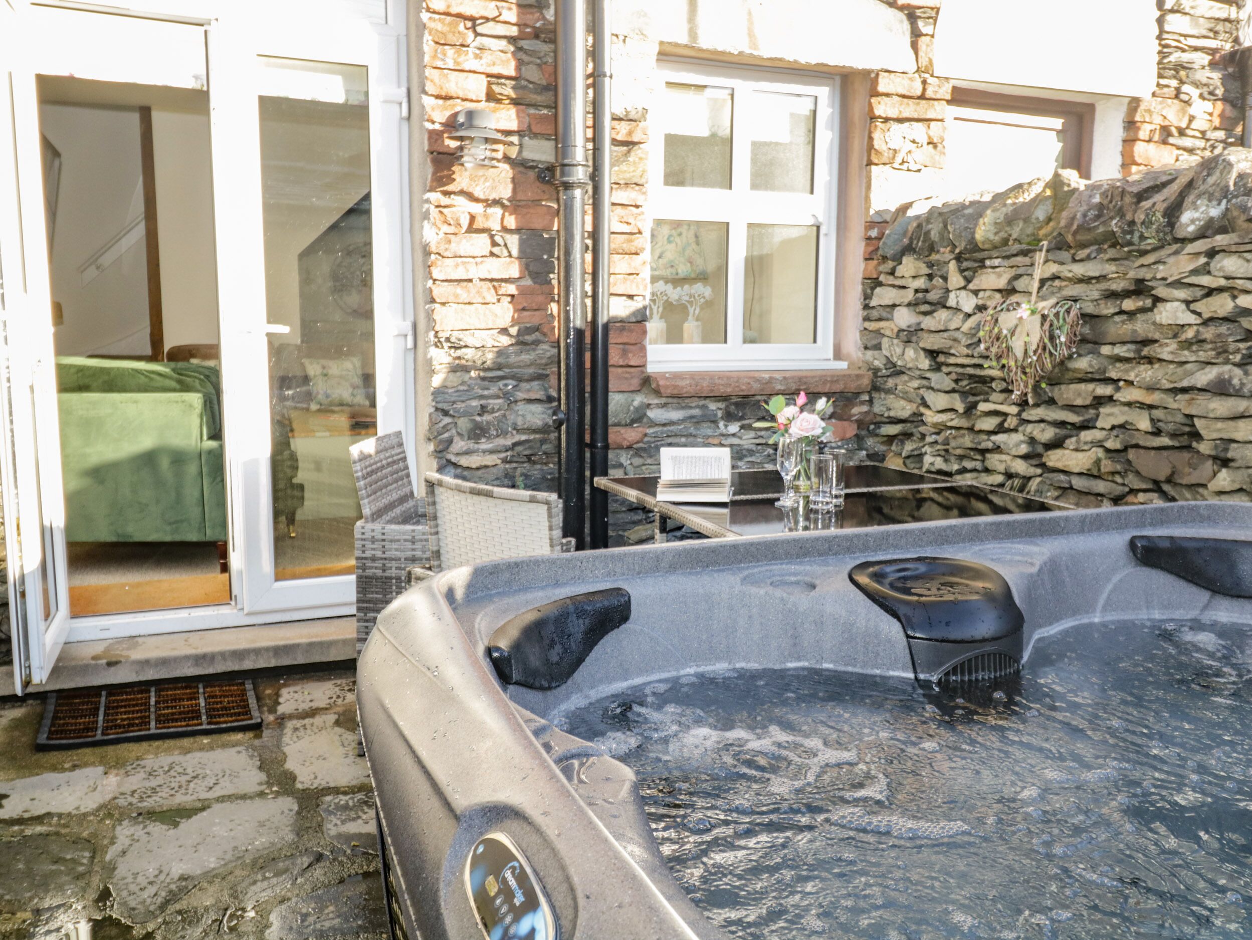 A hot tub in a stone-walled outdoor area with a sliding glass door leading to a room with a green bedspread.
