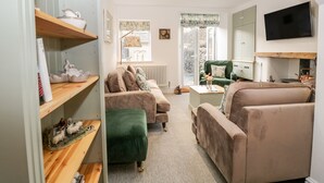 A cozy living room with a sofa, a green armchair, and a coffee table. There is a bookshelf with books and decorative items. A television is mounted on the wall, and a window provides natural light. - Pottergill (Windermere)