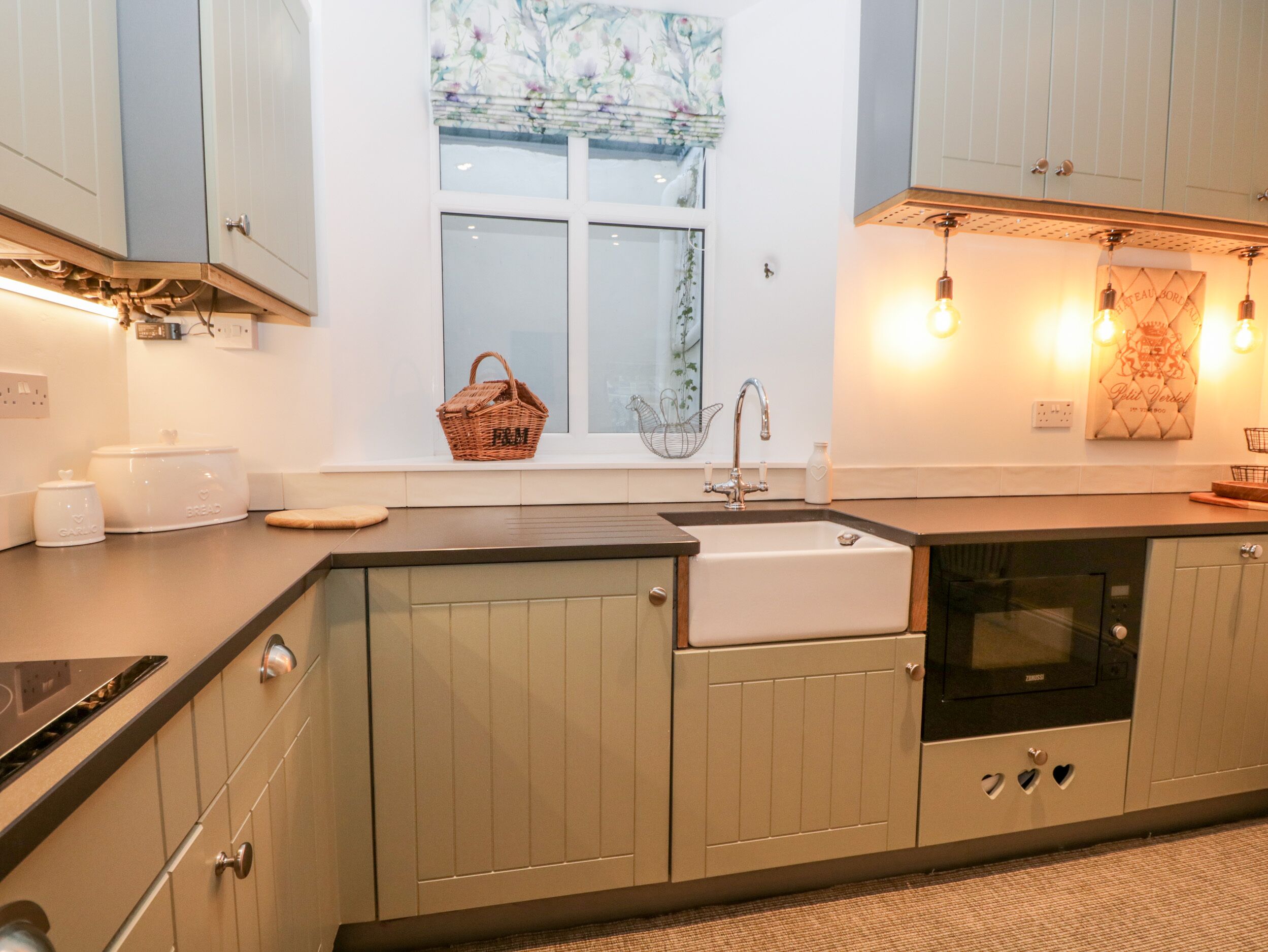 A modern kitchen with light wood cabinets, a built-in oven, a sink, and a window with a floral curtain.