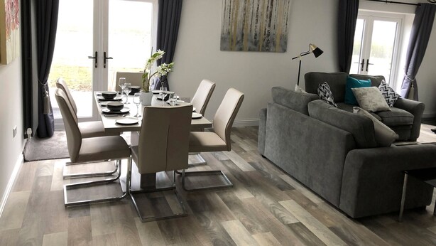 A modern living room with a grey sofa, a dining area with a table set for four, and a large window with curtains. - 2 Barn Cottages (Whitchurch)