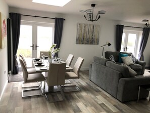A modern living room with a grey sofa, a dining area with a table set for four, and a large window with curtains. - 2 Barn Cottages (Whitchurch)