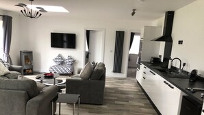 A modern living room with a sofa, a coffee table, and a TV. There is a kitchen area with a stove and cabinets. - 2 Barn Cottages (Whitchurch)