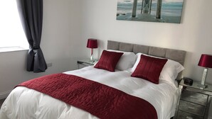 A neatly made bed with a white and red bedspread, two red pillows, and a red throw blanket. There is a painting of a pier on the wall above the bed. - 2 Barn Cottages (Whitchurch)