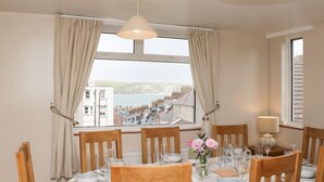 A dining area with a table set for six, a vase with flowers, and a view of a coastal town through large windows. - Seaview (Swanage)