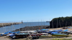 Un puerto deportivo con varias embarcaciones amarradas, un faro al fondo y un acantilado rocoso.