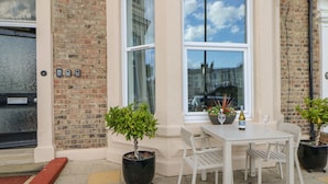Un edificio de ladrillo con una puerta blanca, una ventana con vista al cielo, y una mesa con sillas y plantas en macetas.