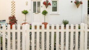 A white house with a white picket fence, a door, and two windows. There are potted plants and hanging flower baskets on the wall. - Love Cottage (Tenby)