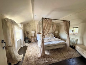 Una cama con dosel y cuatro columnas, un tapete mullido, una mesita de noche con una lámpara y una ventana con vistas.