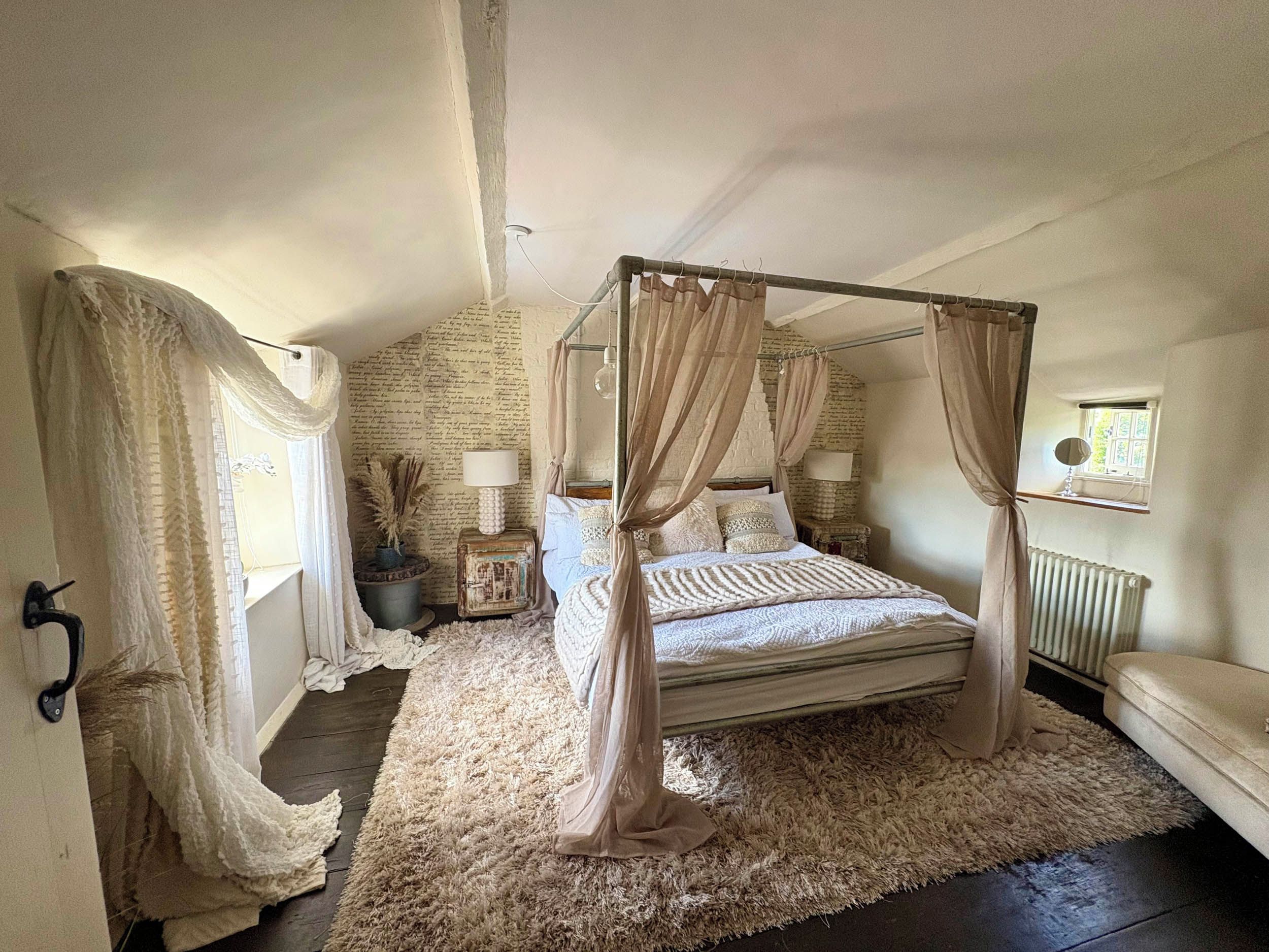 A four-poster canopy bed with a fluffy rug, a small bedside table with a lamp, and a window with a view.