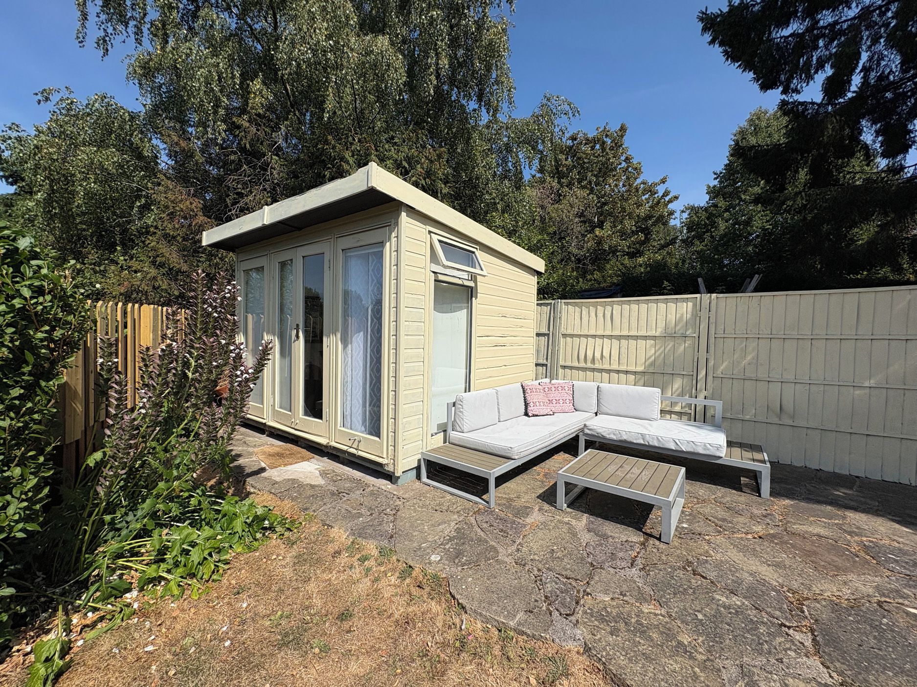 A small, light-colored wooden structure with a sliding door, a white sofa, and a wooden table in a backyard setting.