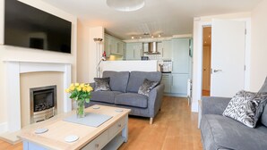 A modern living room with a fireplace, grey sofas, a coffee table, and a kitchen in the background. - 12 Combehaven (Salcombe)