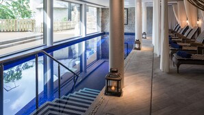 A serene indoor pool area with a clear view of the outside, a set of stairs leading down to the pool, and a row of lounge chairs with cushions. - 12 Combehaven (Salcombe)
