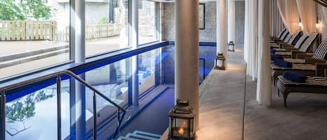 A serene indoor pool area with a clear view of the outside, a set of stairs leading down to the pool, and a row of lounge chairs with cushions.