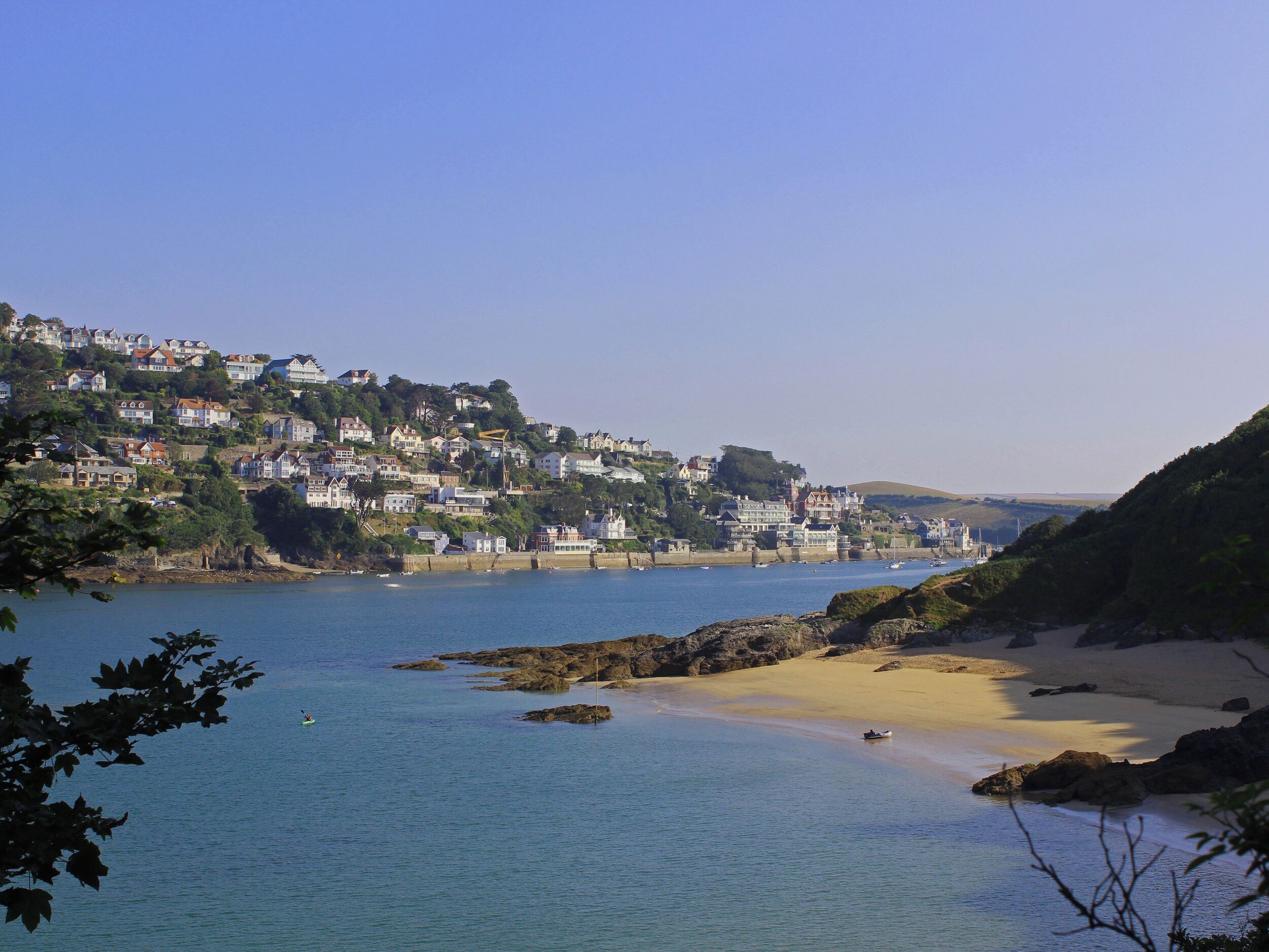 Uma vila costeira com casas numa encosta, uma praia de areia e águas calmas.