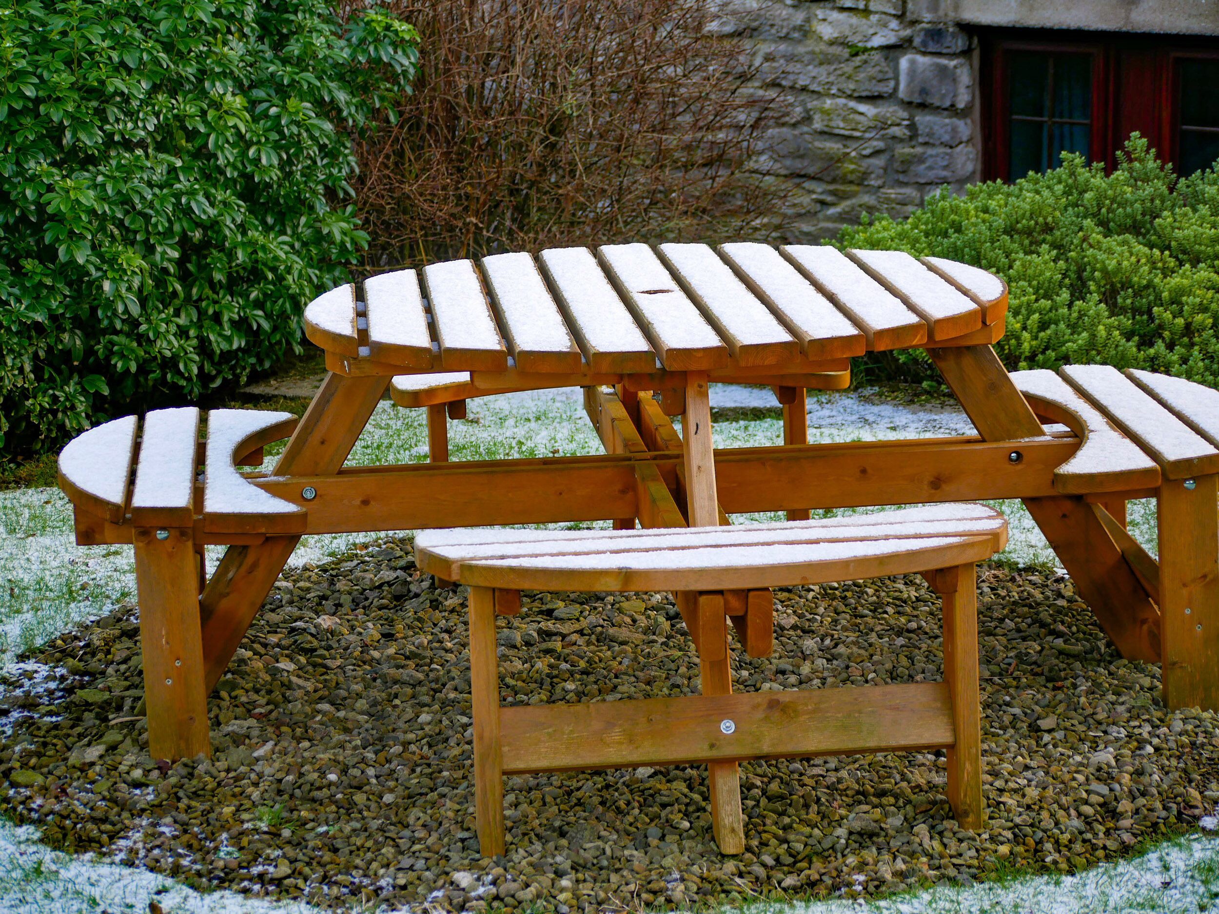 Une table de pique-nique en bois, avec des bancs, posée sur un sol de gravier, avec de la verdure en arrière-plan.