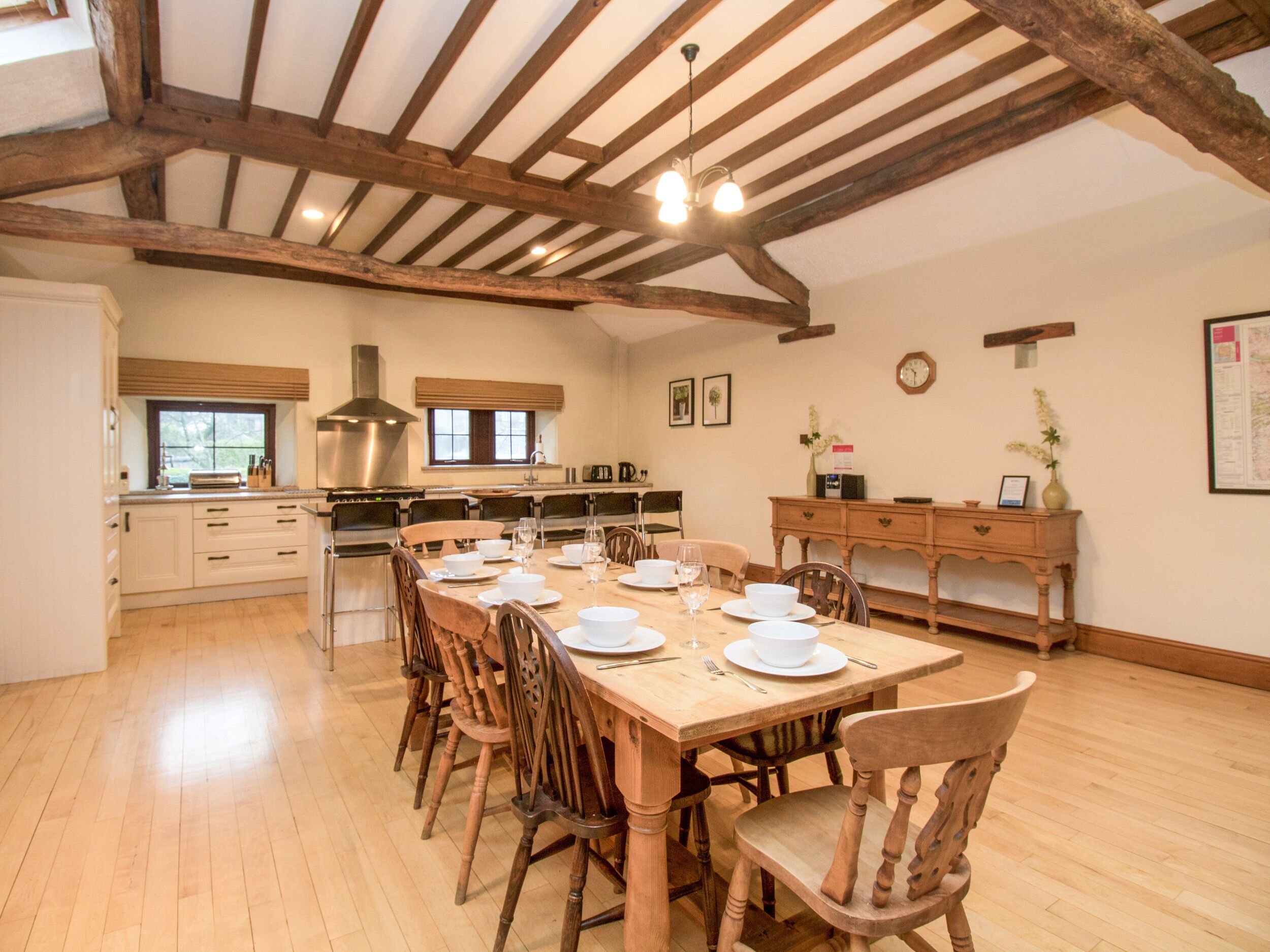 A spacious dining area with a wooden table set for six, surrounded by wooden chairs. A kitchen with modern appliances and a large window is visible in the background.