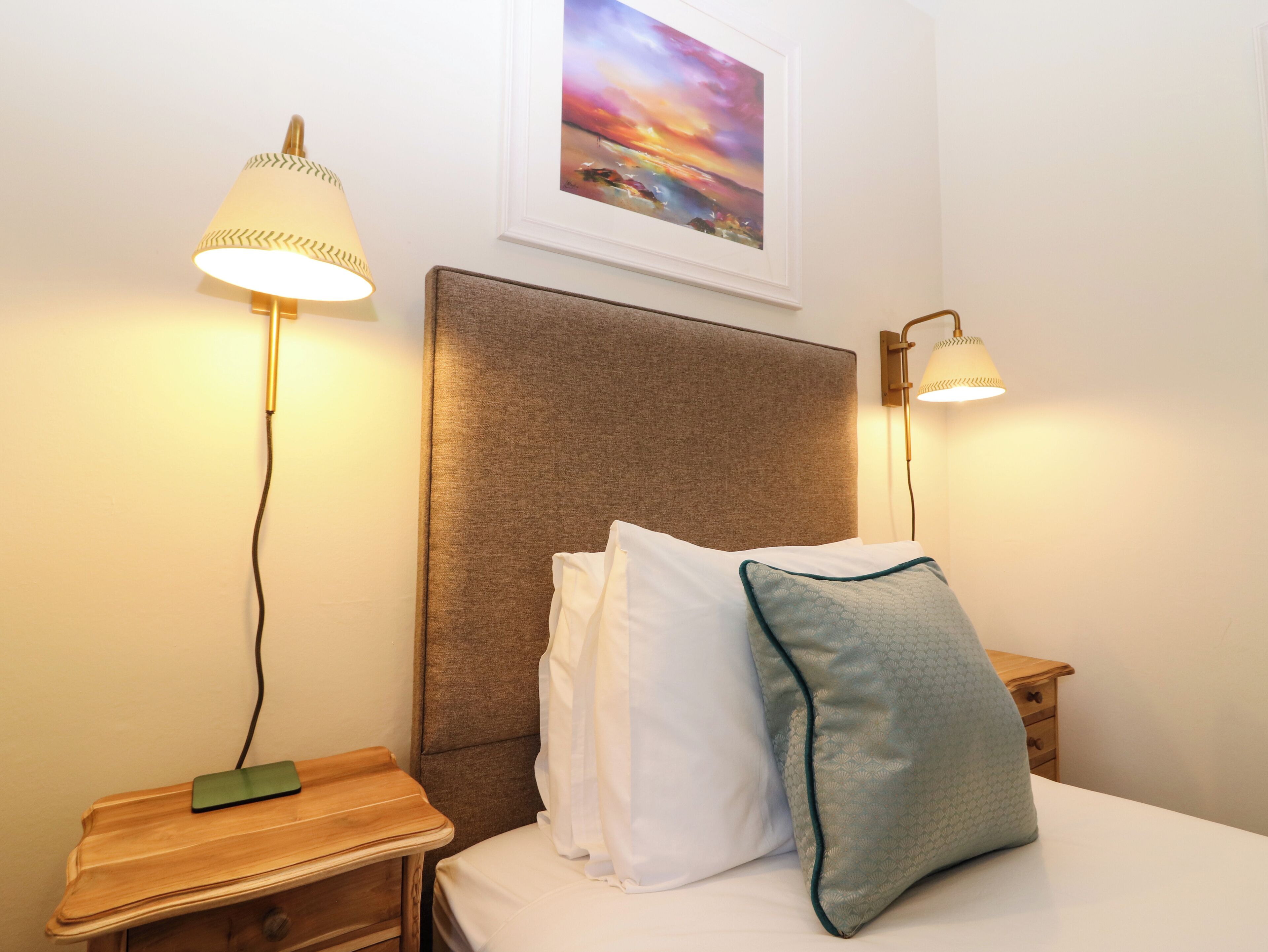 Una cama bien hecha con sábanas blancas y dos almohadas, flanqueada por dos mesitas de noche con lámparas. Se observa una obra de arte enmarcada en la pared sobre la cama.