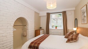 A bedroom with a white brick wall, a bed with white bedding and plaid pillows, a hanging light fixture, a window with curtains, and a small table with a plant. - The Lanterns (Buxton)