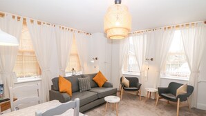 A modern living room with a grey sofa, two armchairs, a coffee table, and a dining table with chairs. There is a large fluffy rug, white curtains, and a hanging light fixture. - The Lanterns (Buxton)
