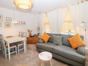 A living room with a grey sofa, two orange cushions, a white coffee table, a dining table with chairs, and a wooden side table with a lamp. - The Lanterns (Buxton)