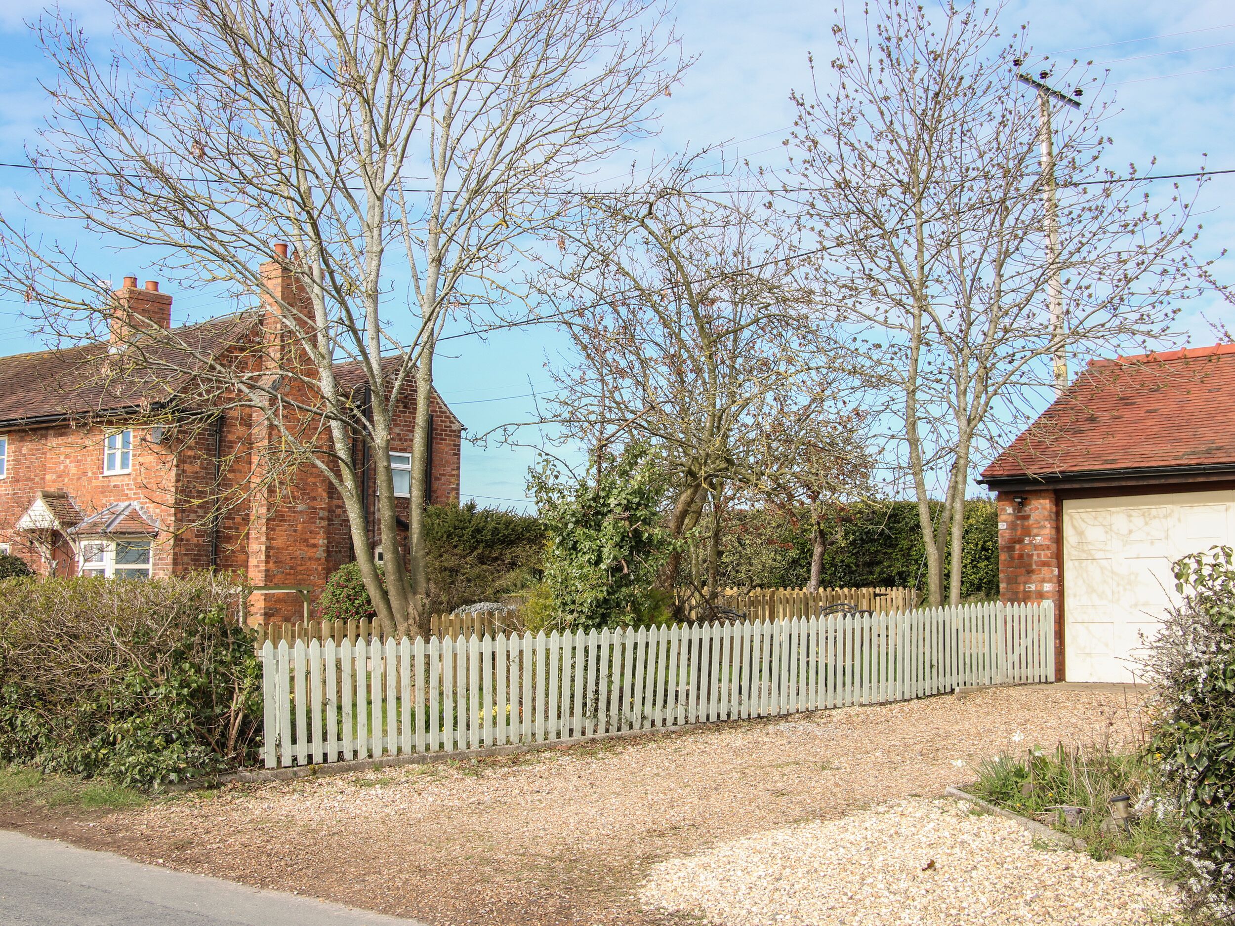 A residential area with a gravel driveway, a white picket fence, and a brick house with a garage.