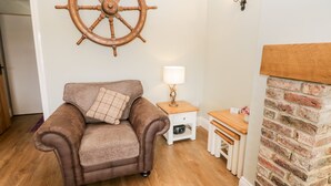 A cozy living room with a brown leather sofa, a wooden side table with a lamp, a wall-mounted ship's wheel, and a brick fireplace. - Shipswheel Cottage at Broadings Farm (Whitby)