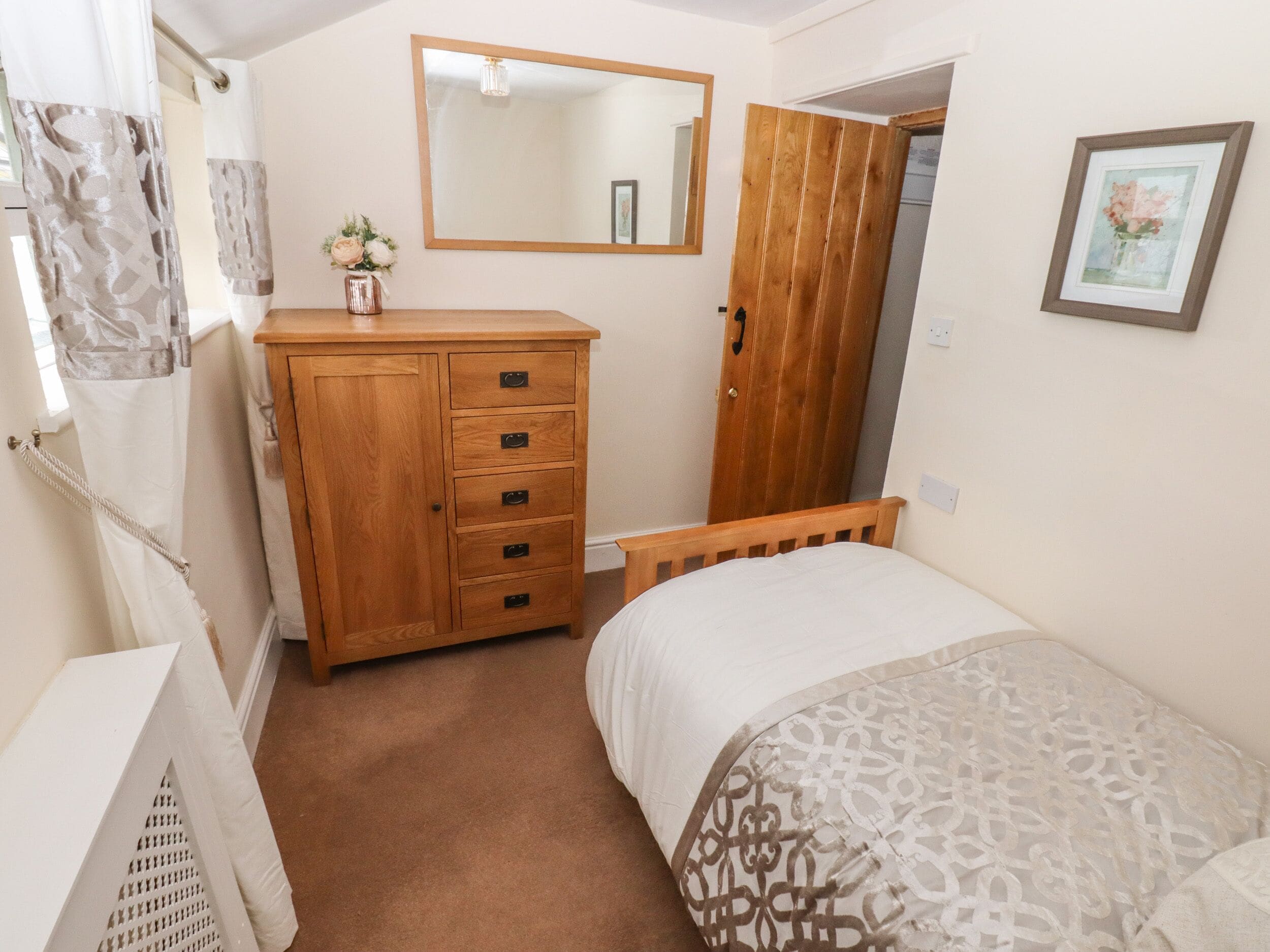 Une chambre à coucher avec une commode en bois, un lit avec une literie à motifs, un miroir et une porte donnant sur une autre pièce.