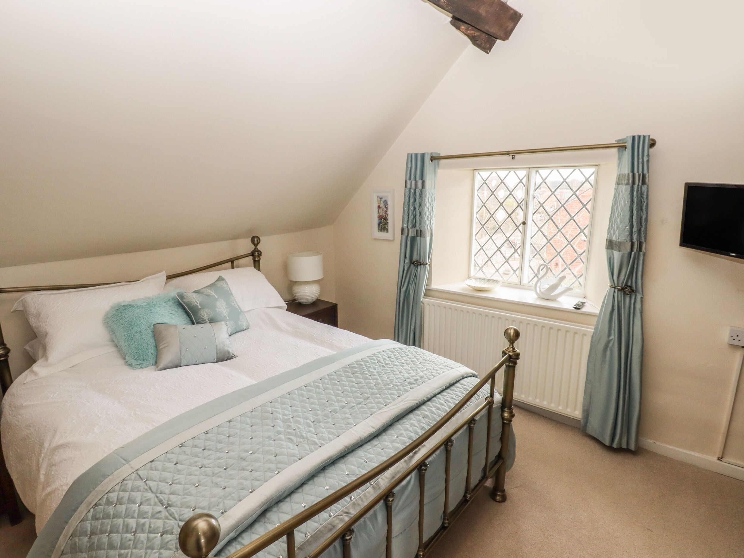 A bedroom with a bed, a bedside table, a window with curtains, and a wall-mounted TV.
