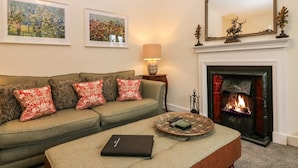 Una sala de estar con un sofá verde, cojines estampados en rojo y blanco, un reposapiés negro, una chimenea y dos cuadros con motivos florales.