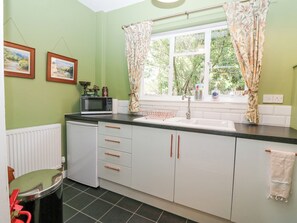 Une cuisine équipée d’un four à micro-ondes, d’un évier et de placards. Deux tableaux encadrés ornent le mur, et une fenêtre est agrémentée de rideaux.