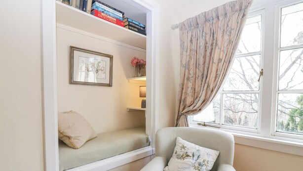 Una habitación acogedora con un asiento empotrado junto a la ventana, un sillón con un cojín estampado y una estantería llena de libros.