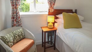 A bedroom with a bed, a bedside table with a lamp, a chair, and curtains. - Branklyn Garden Cottage (Perth)