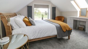 A bedroom with a sloped ceiling, a bed with white bedding and a grey throw, a wooden headboard, a bedside table with a lamp, a chair, a desk with a mirror, and a window with curtains. - Pheasant's Roost (Hull)