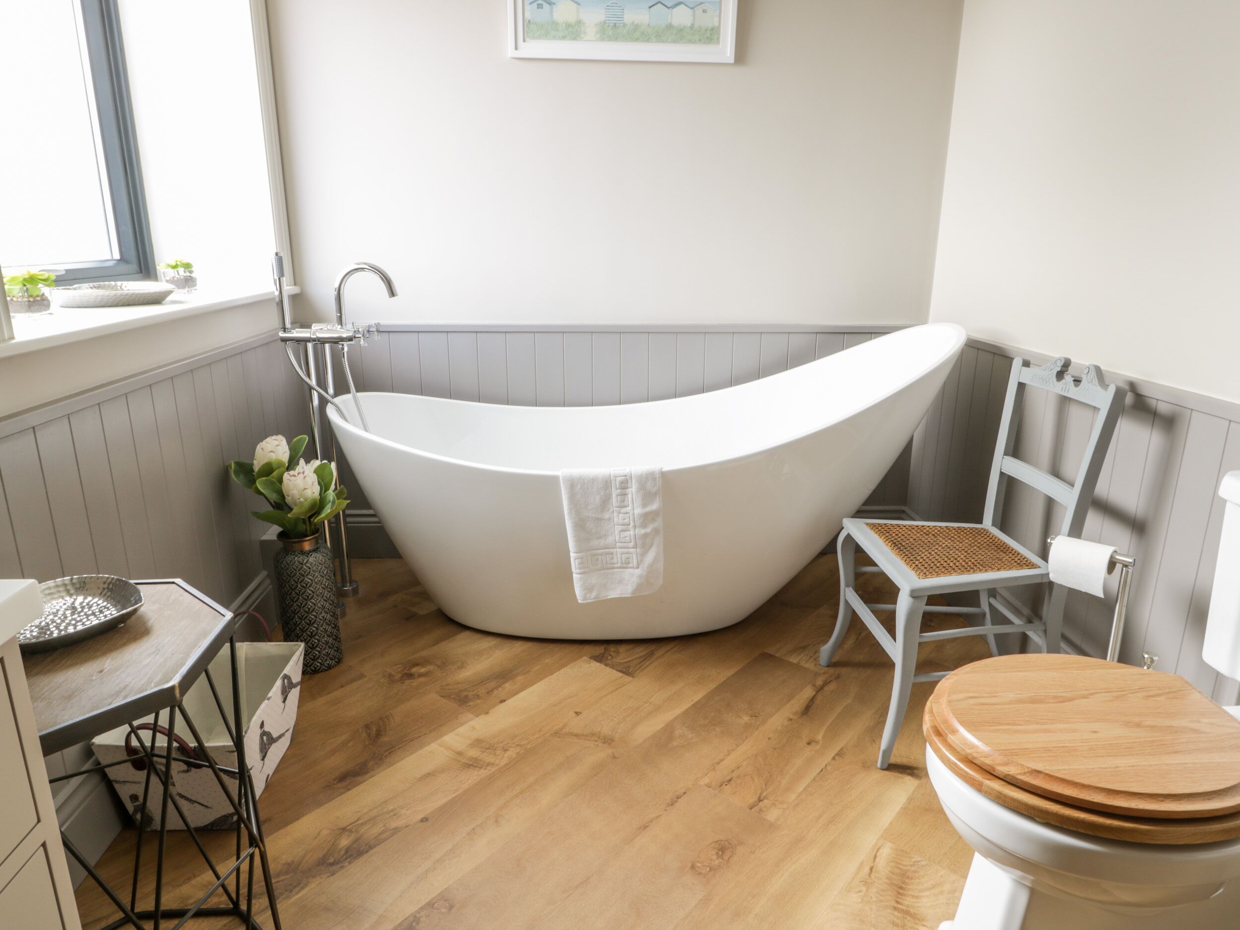 A modern bathroom with a freestanding bathtub, a wooden toilet, and a small table with a vase of flowers.