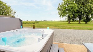 An outdoor hot tub with a step leading into it, surrounded by a gravel area and a grassy field with trees in the background. - Pheasant's Roost (Hull)
